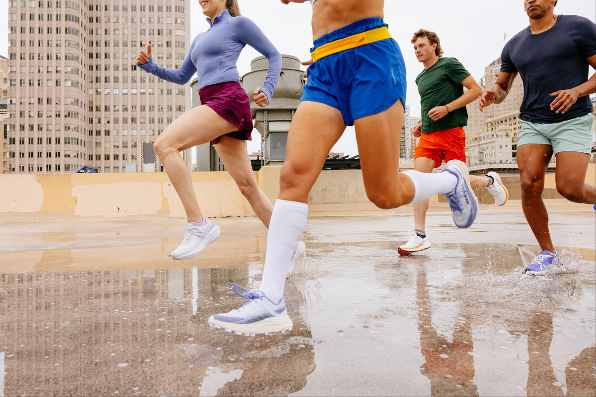Four runners jogging on a wet city rooftop with buildings in the background. The runners are shown from the waist down, wearing athletic apparel in blue, purple, green, and navy, with white socks and running shoes.